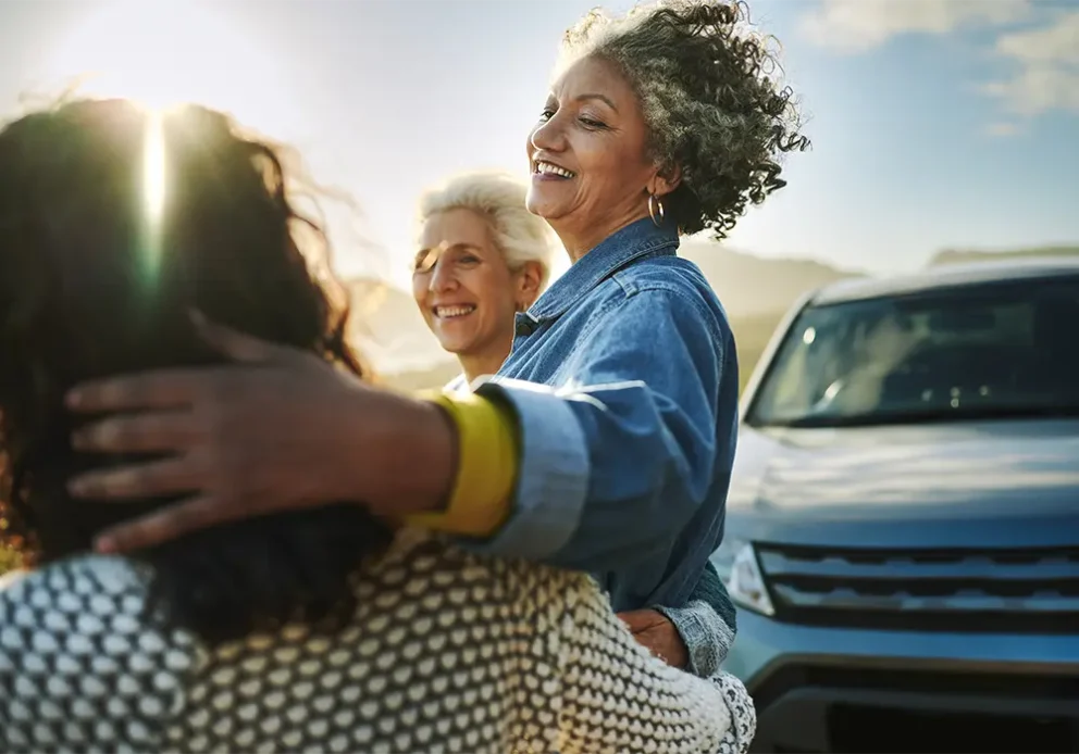 Carefree group of mature female friends standing arm in arm by their car parked on a road running along a scenic coastline.