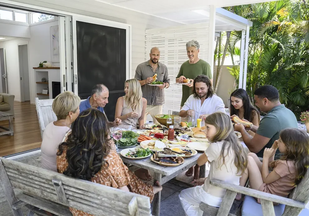 Multi-generation family seated and standing around outdoor patio table, eating barbecued meat, salad, cheeses, and fruit, smiling and talking.