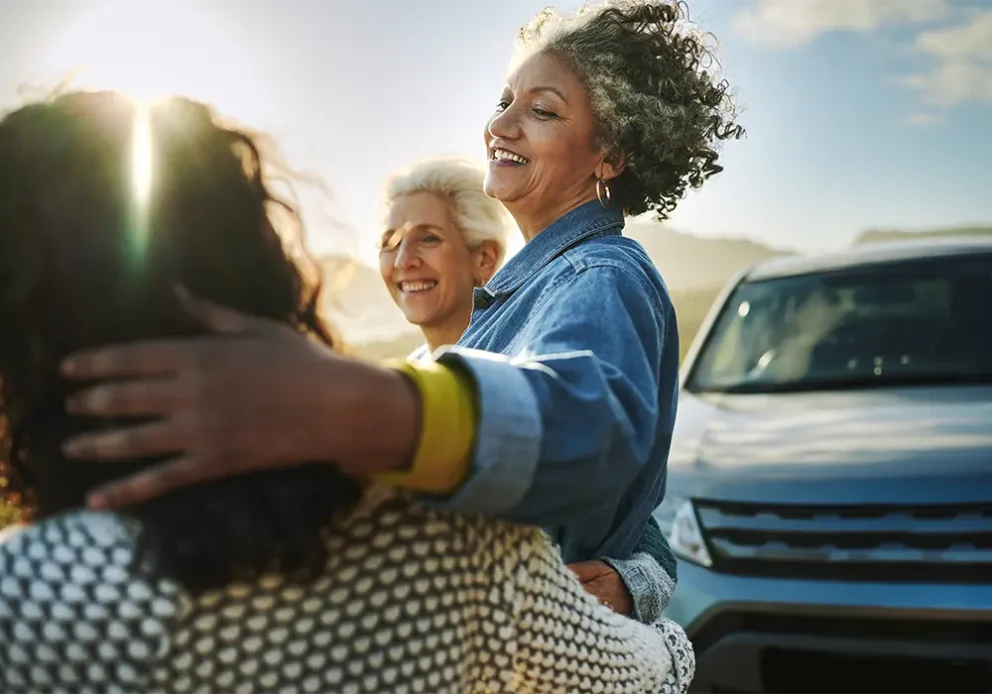 Carefree group of mature female friends standing arm in arm by their car parked on a road running along a scenic coastline.